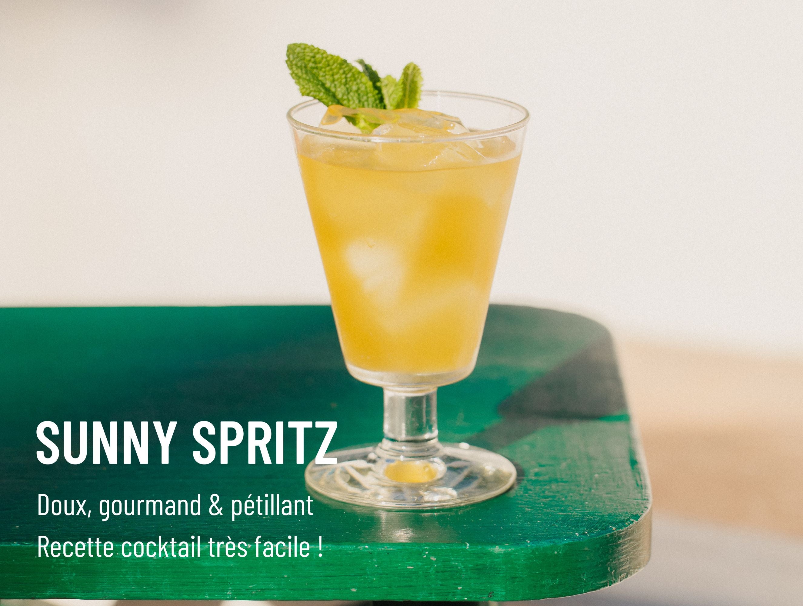 Verre de Sunny Spritz, cocktail jaune vif décoré de menthe, posé sur une table verte en extérieur
